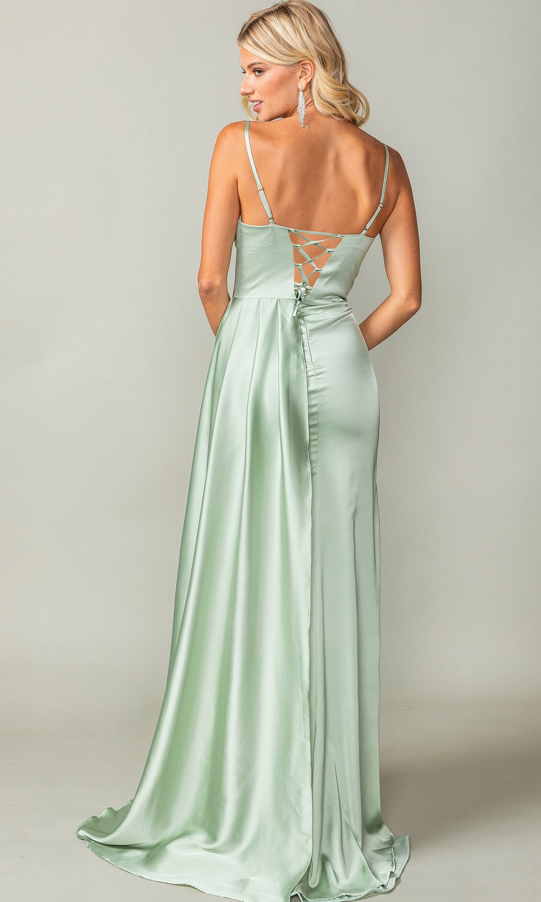A woman stands facing away from the camera in the Beaded-Bust Long Prom Dress: Dancing Queen 4412, a light green gown with thin straps and lace-up back detail, against a plain background.
